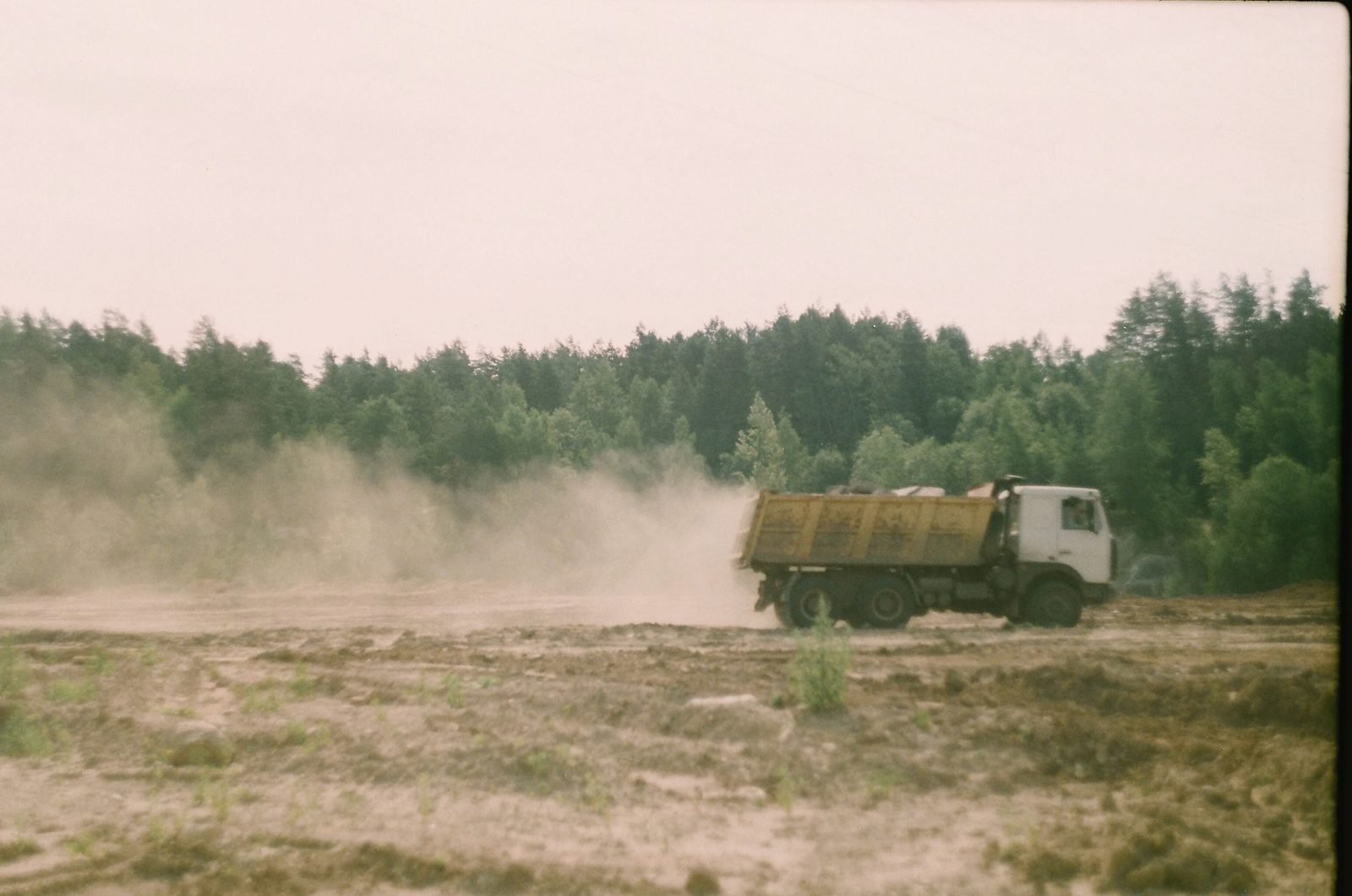 Dump truck crossing a dusty site with woods behind